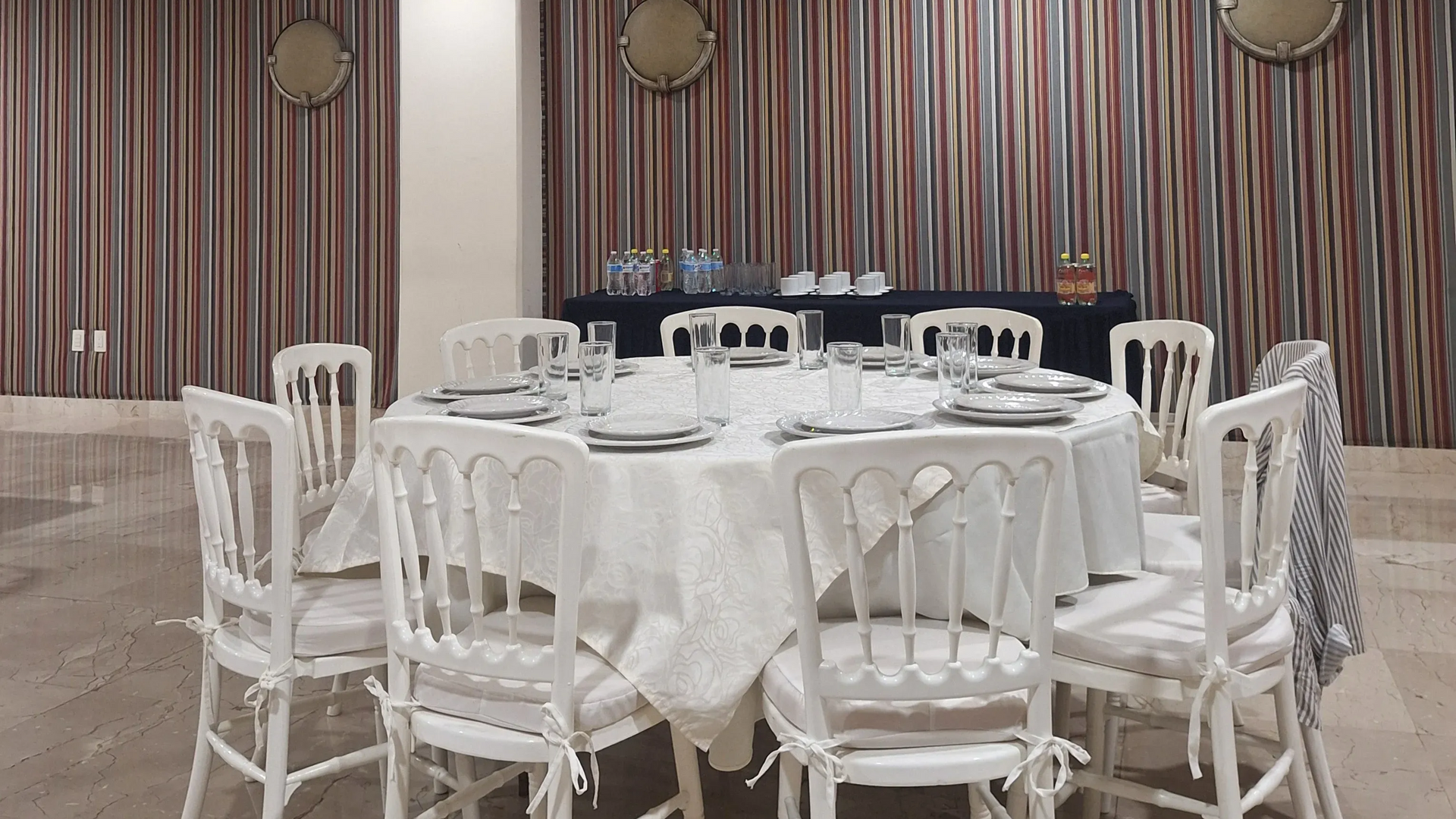 Una mesa redonda con mantel blanco y sillas blancas en una habitación con cortinas a rayas y un aparador al fondo.