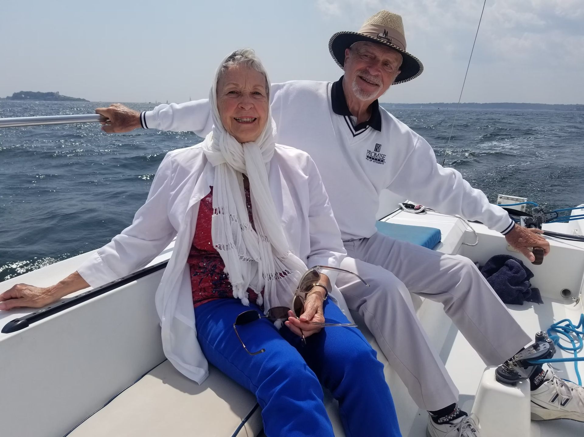 A smiling pair sits together on a sailboat on a sunny day, wearing nautical attire, with the ocean in the background.