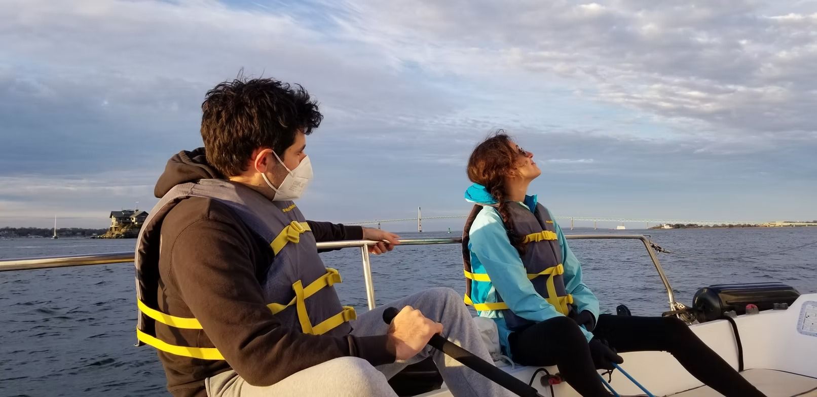Two people wearing life vests on a small boat at sea, looking toward the horizon during sunset.