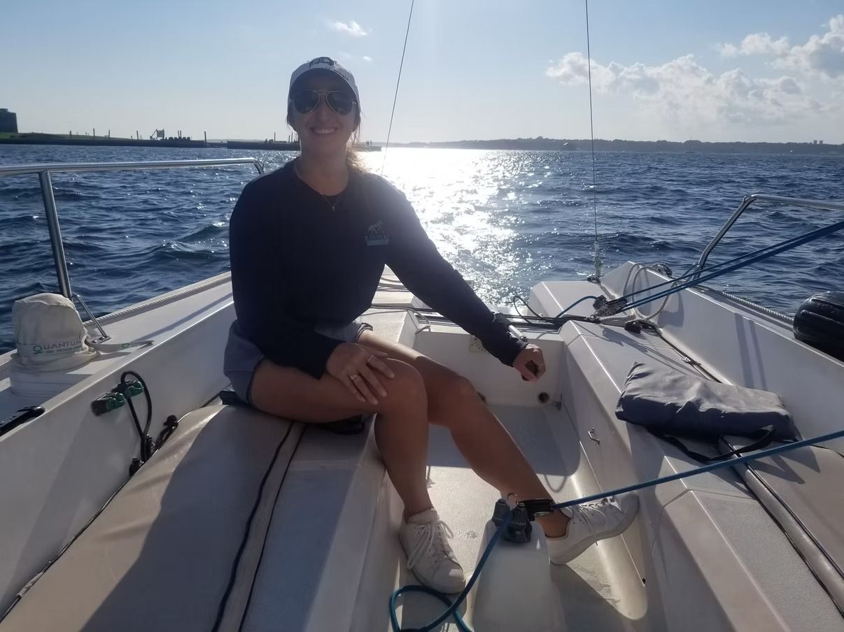 A person wearing a hat and sunglasses sits on a sailboat on a sunny day with the ocean and sky in the background.