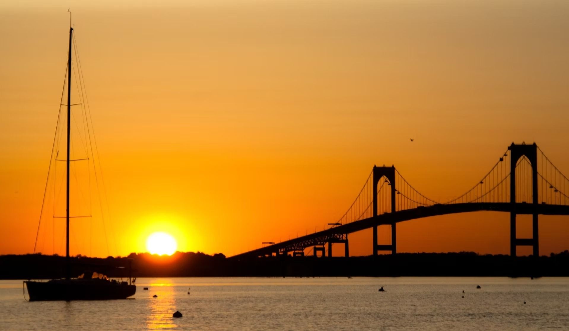 A sailboat silhouette anchored in calm water near a large bridge at sunset with a vibrant orange sky.