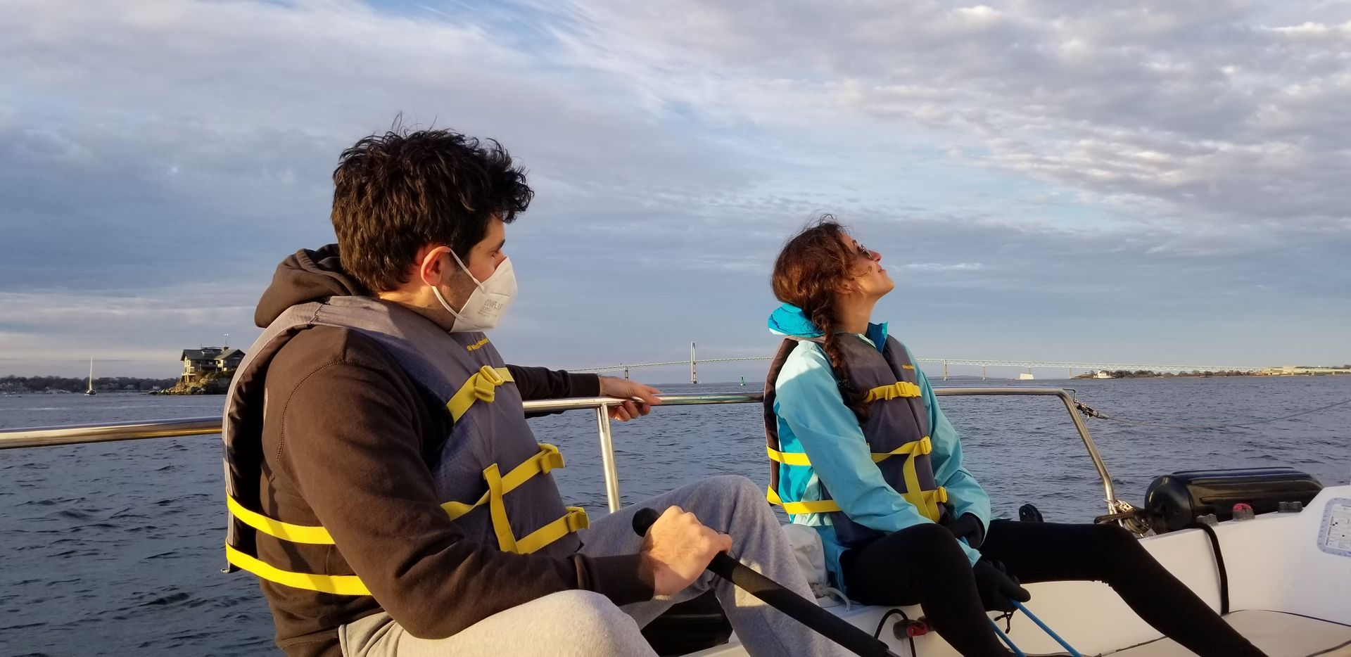 Two people in life jackets ride in a small boat on the water during sunset, looking up at the sky.