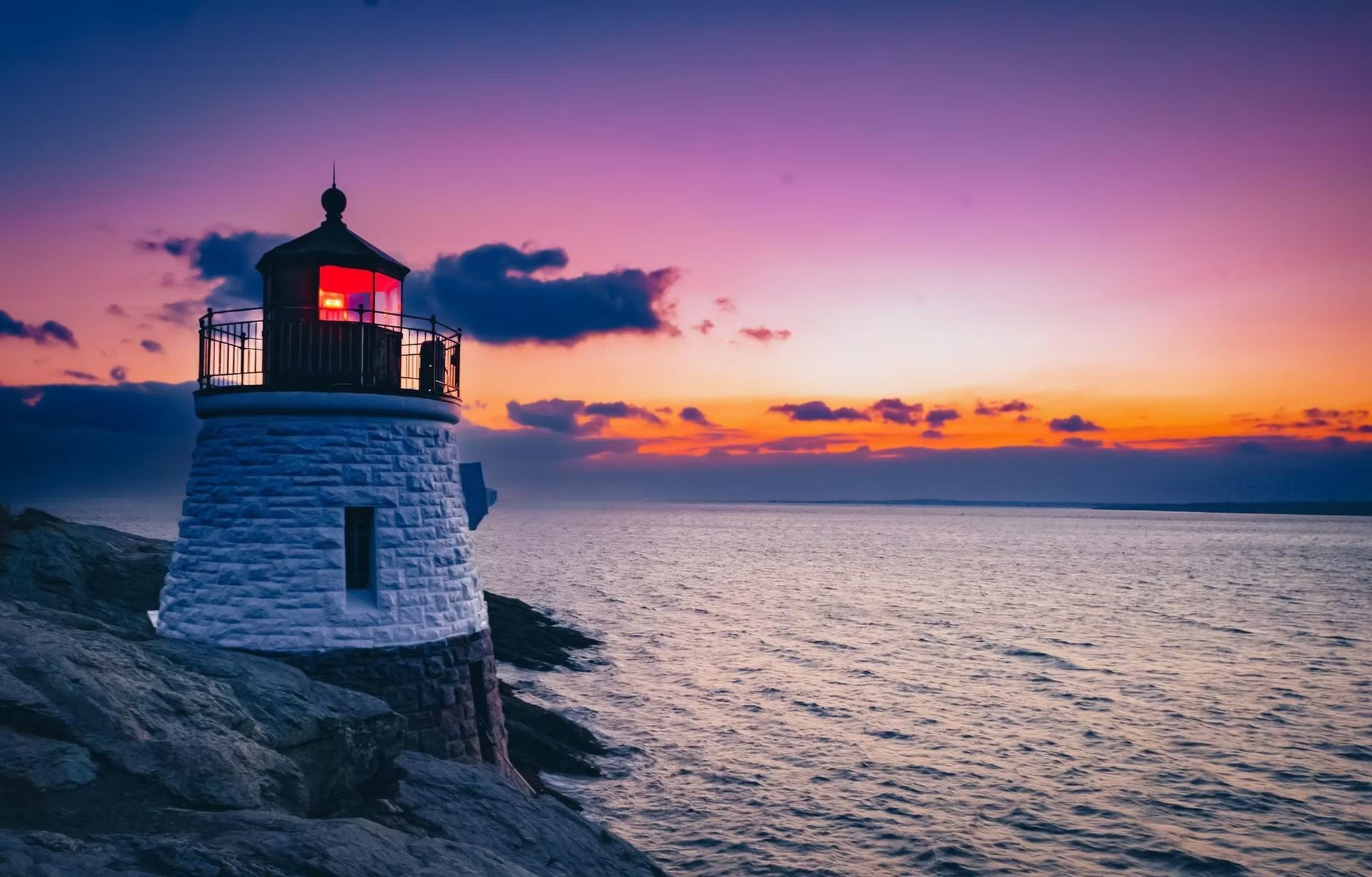 A white stone lighthouse stands on a rocky coast against a vibrant purple and orange sunset over the ocean.
