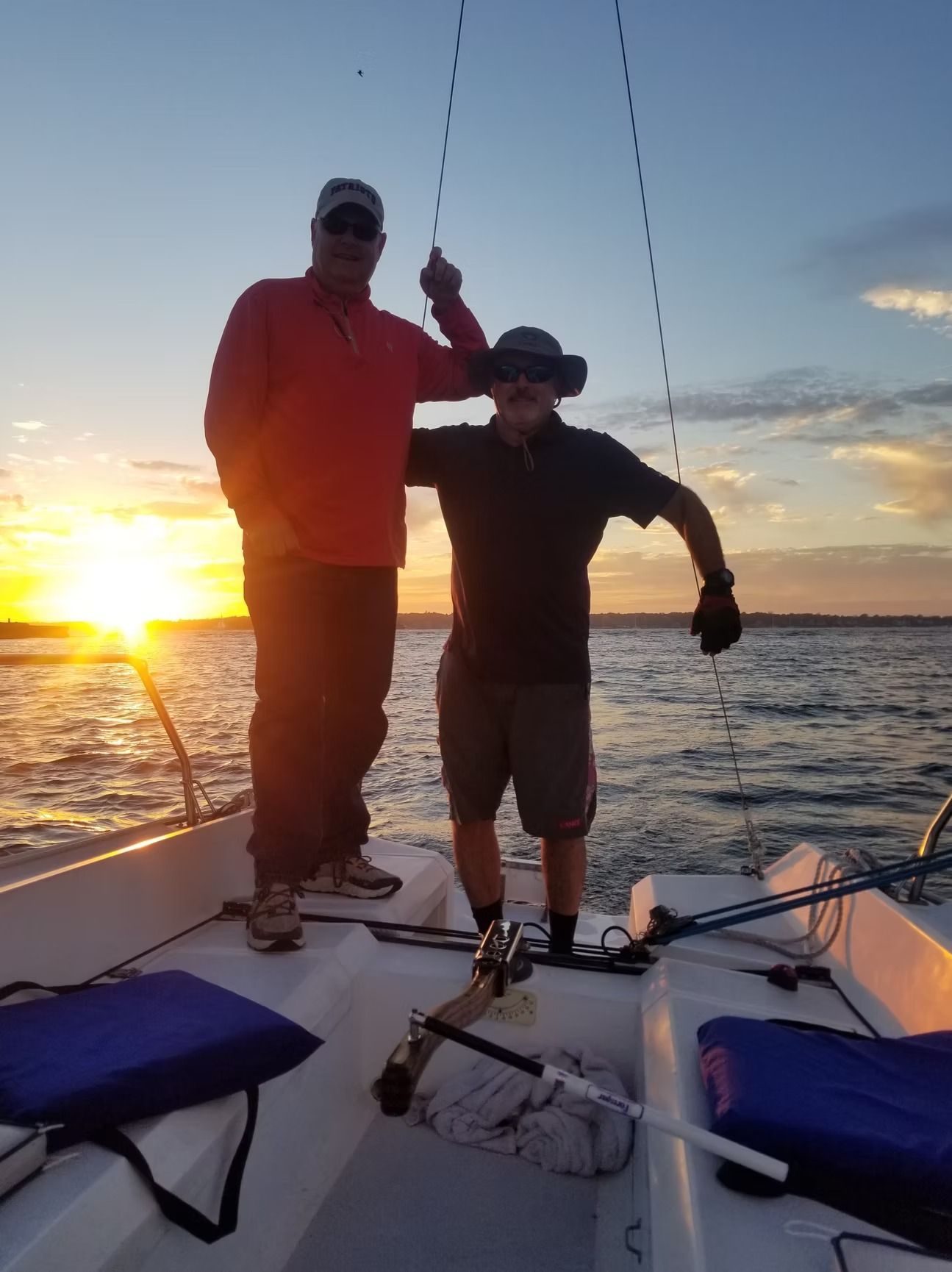 Two people stand on a sailboat during sunset, one in a red shirt and one in a dark shirt, holding onto rigging.