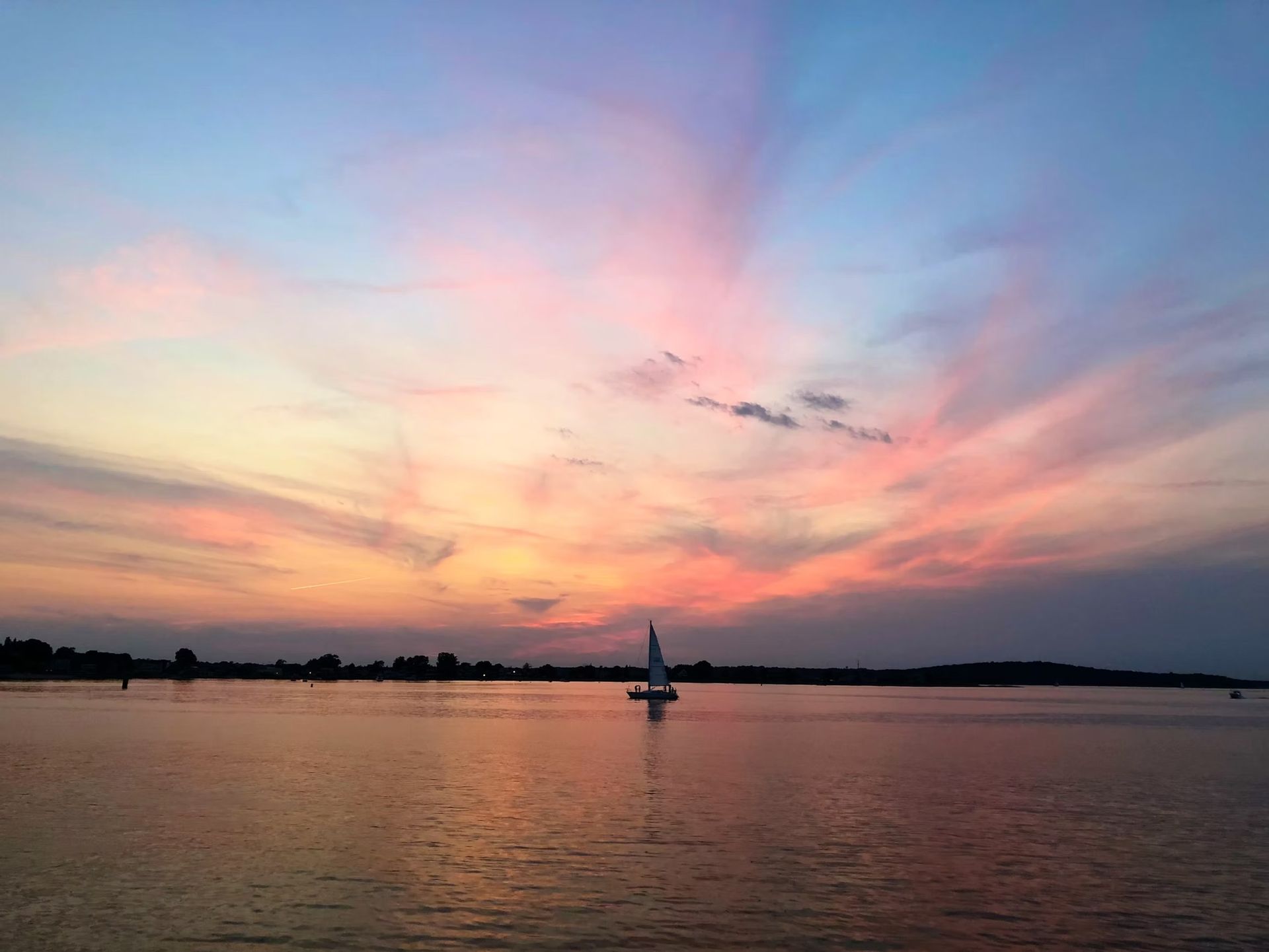 A lone sailboat drifts on a calm lake under a vibrant sunset sky filled with pink, orange, and blue streaked clouds.
