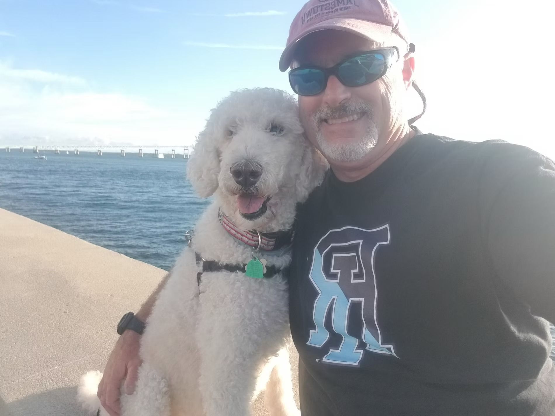 A person wearing sunglasses and a baseball cap poses for a selfie with a smiling white dog near a body of water.