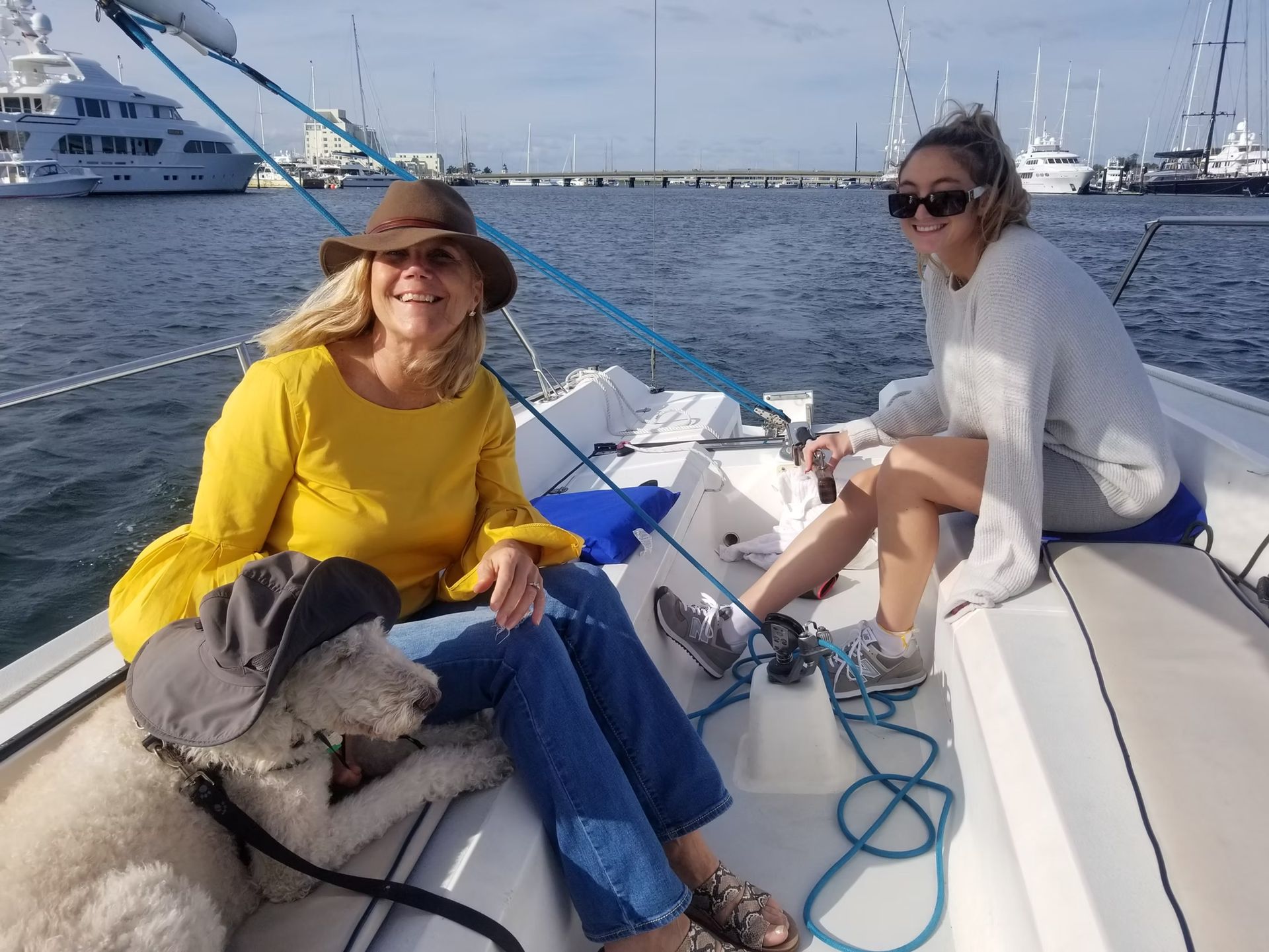Two people and a dog sitting on a sailboat on a sunny day at a marina.