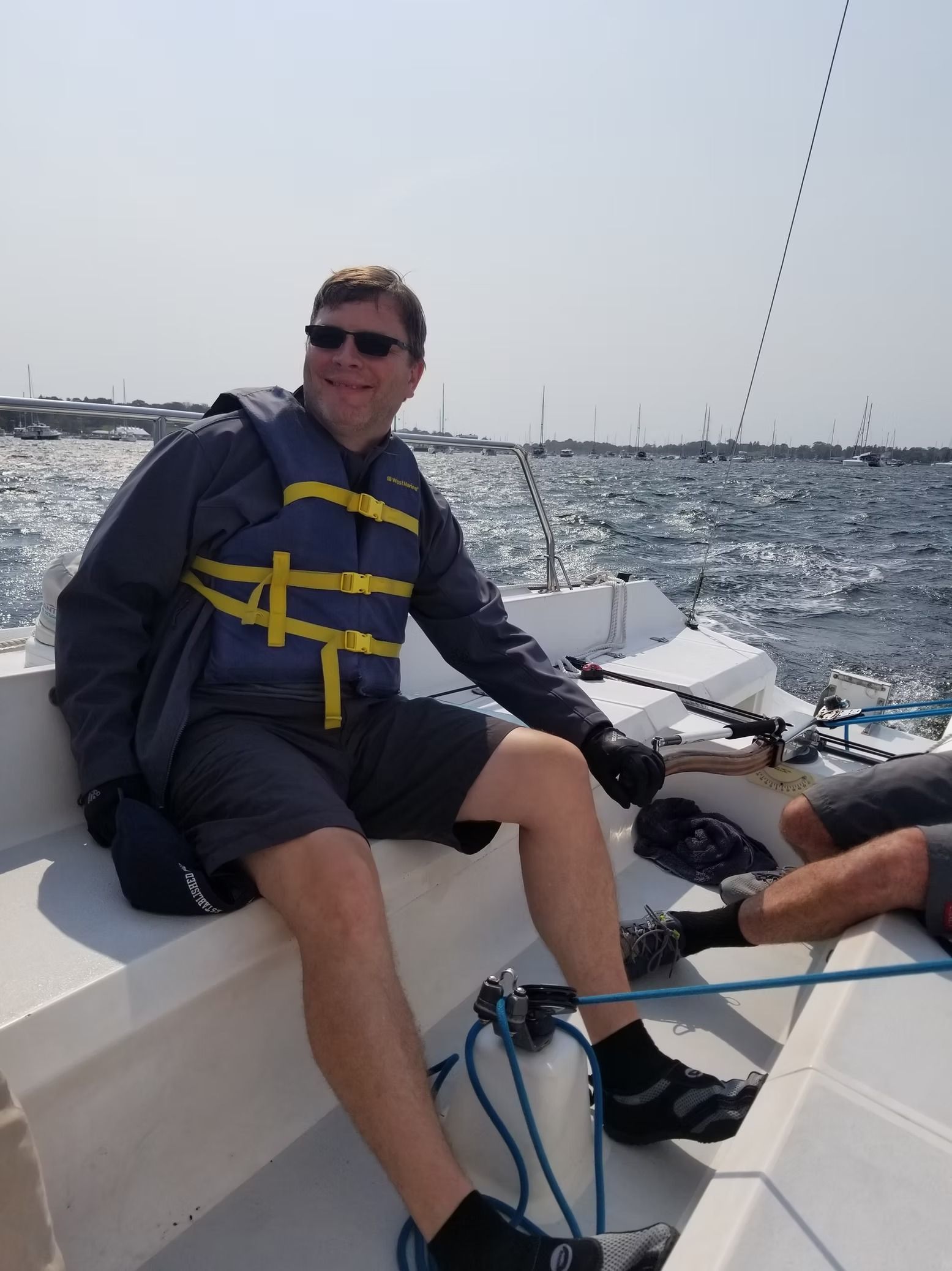 A smiling person wearing a blue life vest and sunglasses sits on a sailboat on the water.
