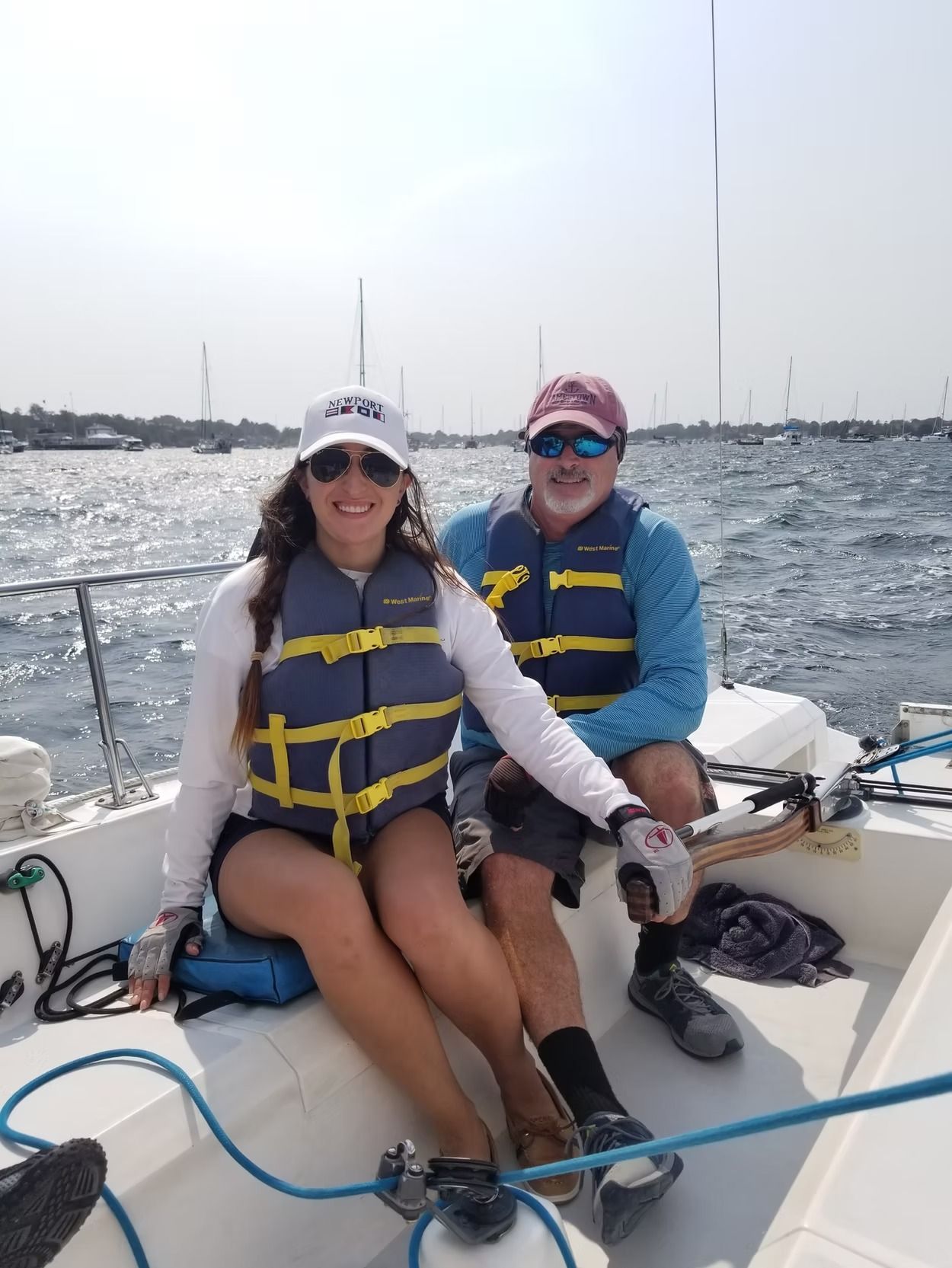 Two people wearing life jackets and hats smile while sitting on a sailboat on the water.