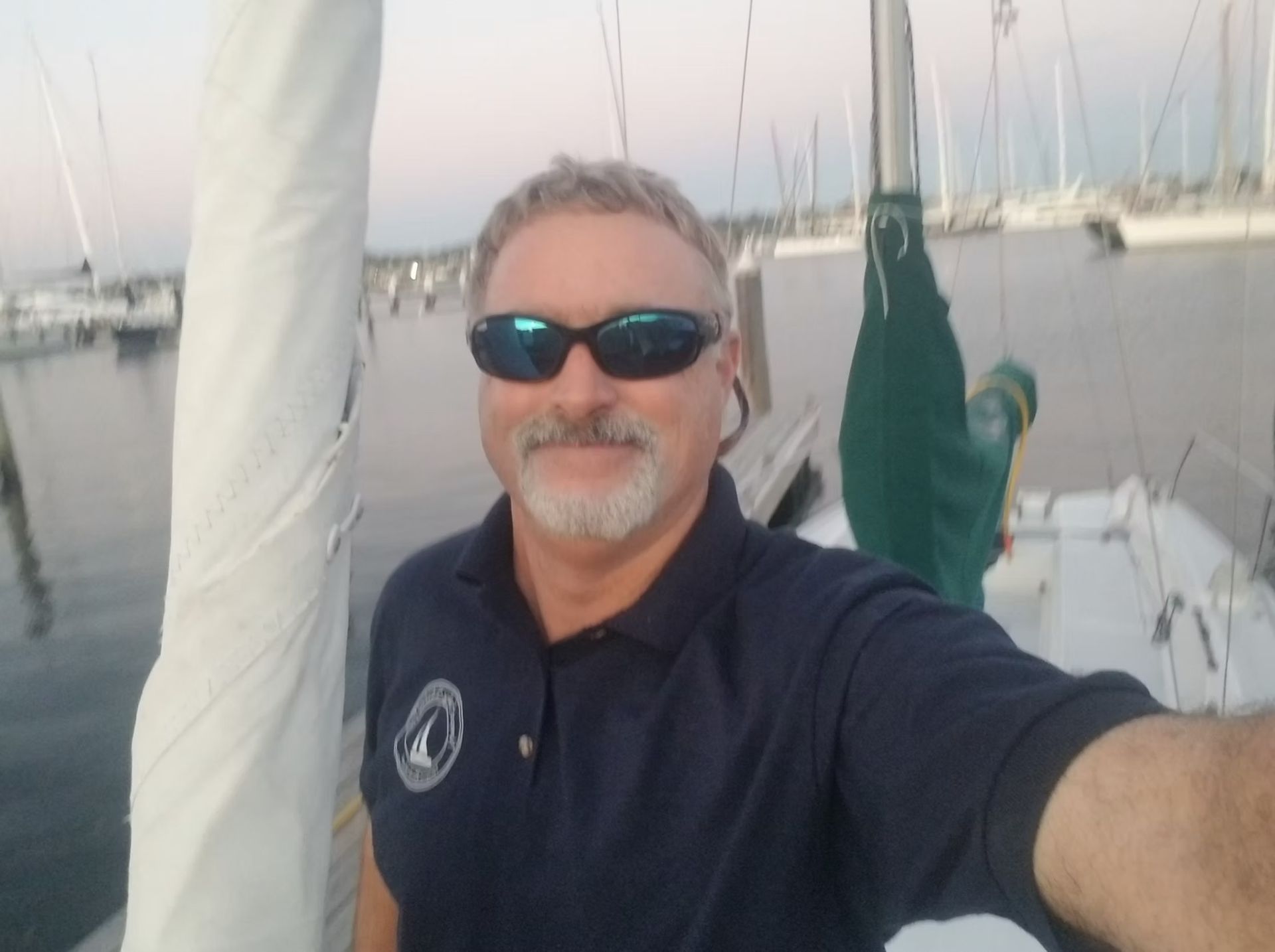 A person with a goatee wearing sunglasses and a navy polo shirt takes a selfie on a sailboat in a marina at dusk.
