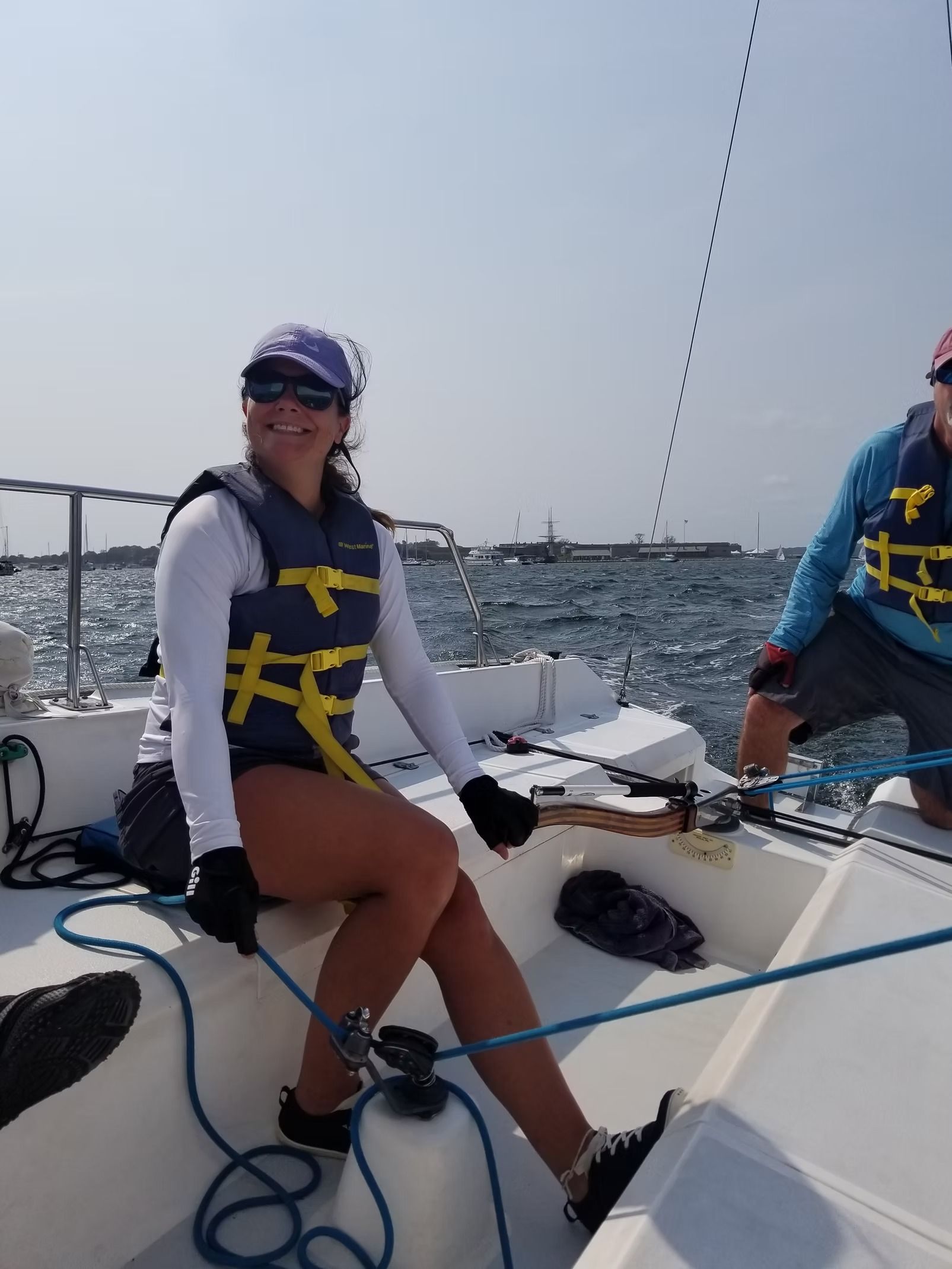 Two people wearing life jackets sit on a sailboat on the water on a sunny day.