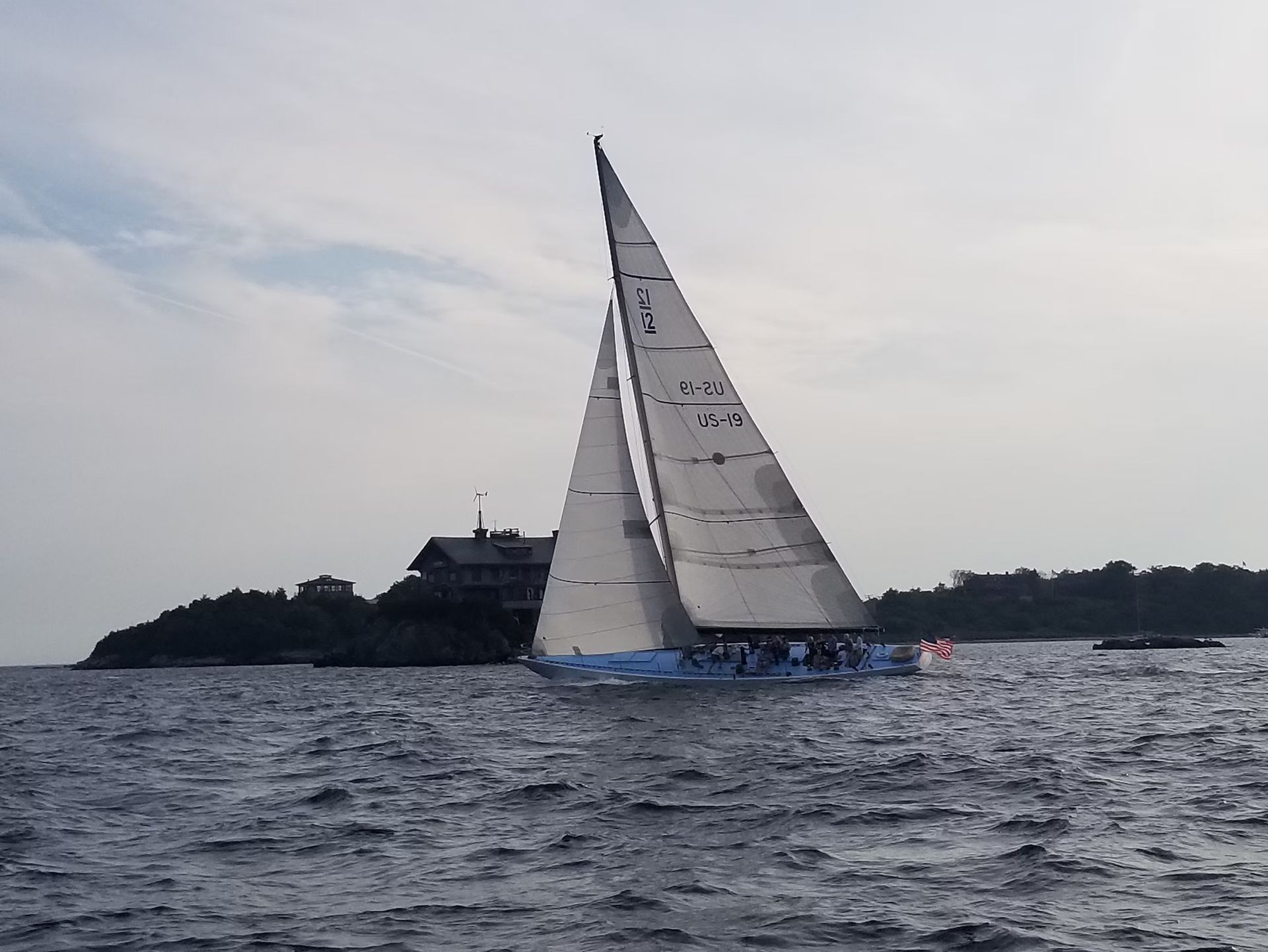 A light blue sailboat with a large white sail navigating near a small, rocky island with a house under a cloudy sky.