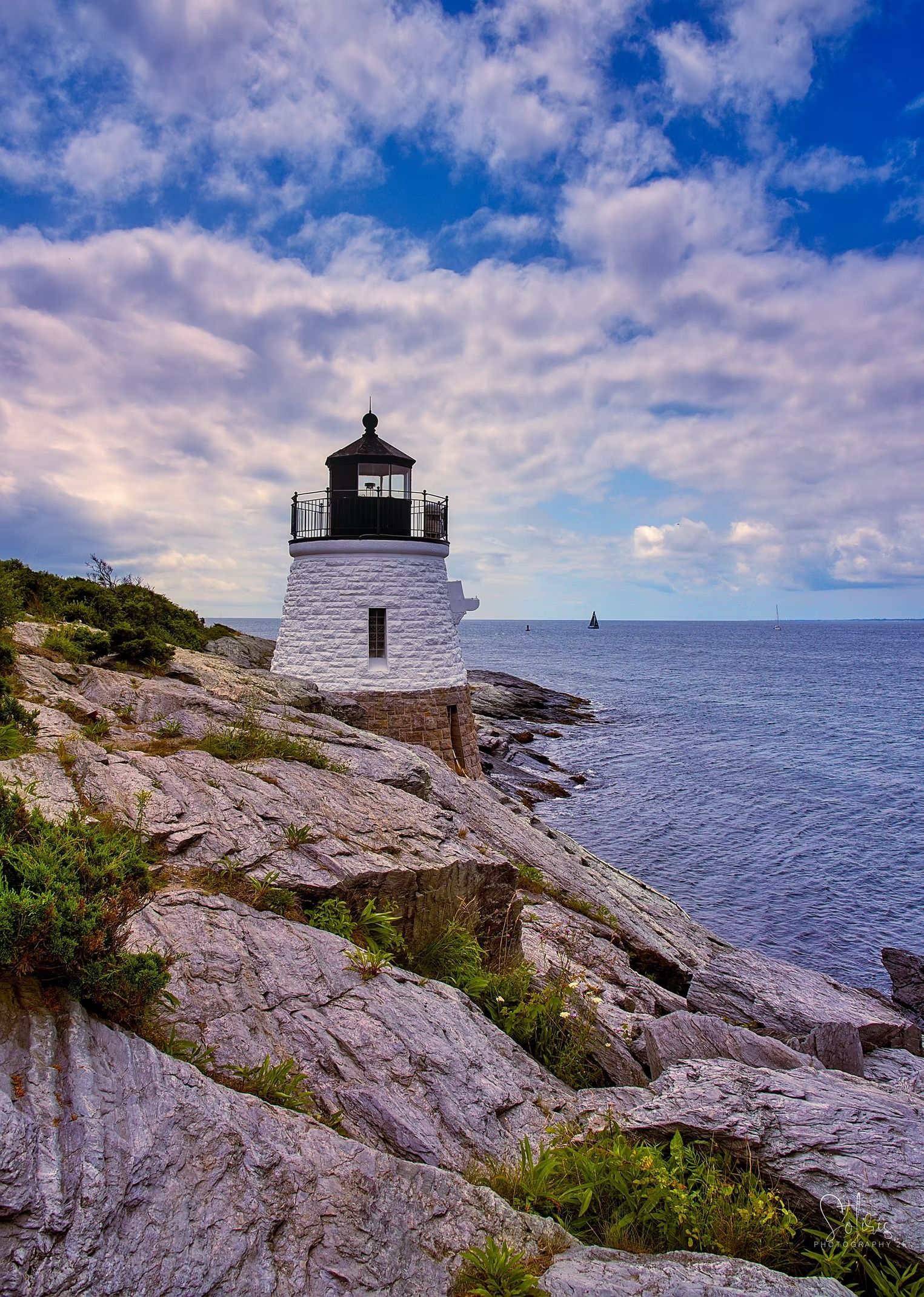 A white lighthouse stands on a rocky, green-veined cliff edge overlooking a vast, dark blue sea under a cloudy sky.