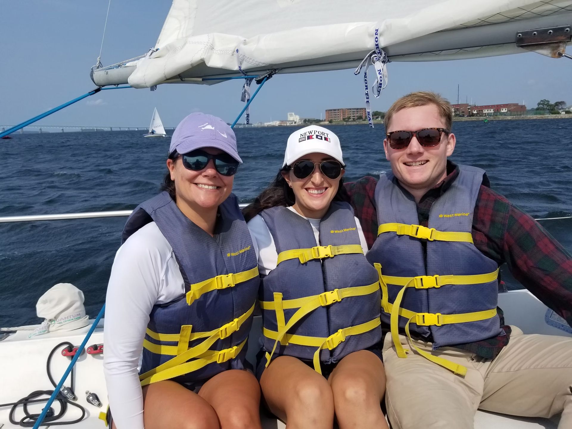 Three smiling people wearing life vests and hats sit on a sailboat on the water during a sunny day.
