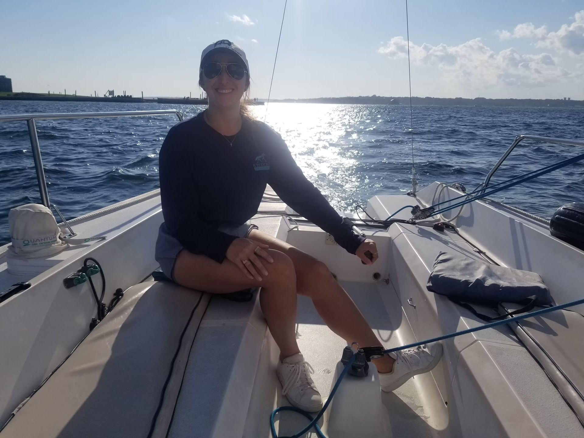 A person in a dark long-sleeved shirt and cap sits on the deck of a sailboat on open water under a bright sky.