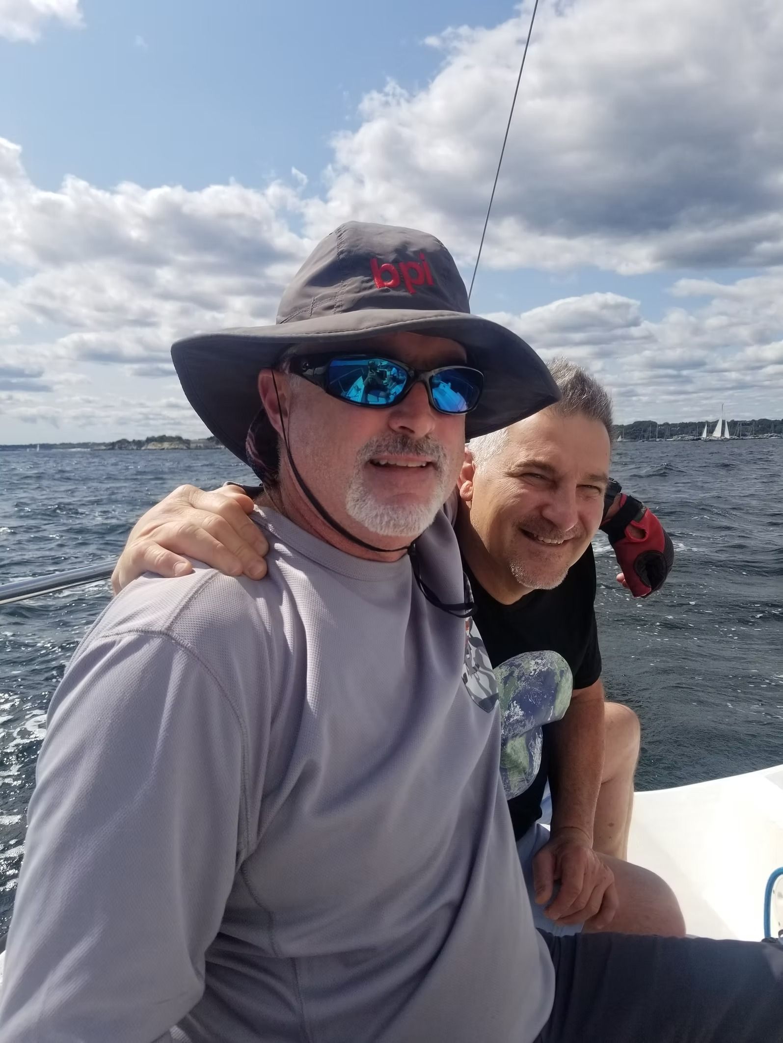 Two men smiling on a boat under a cloudy sky; one wears a sun hat and blue sunglasses, the other has his hand on his back.