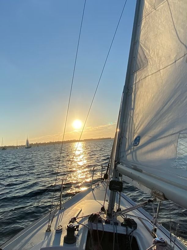 A sailboat deck looking out toward the water at sunset, with a white sail illuminated by golden light.
