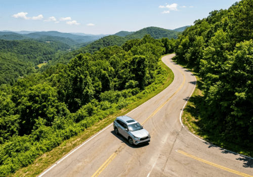 Silver SUV driving along a winding mountain road overlooking lush forested hills on a clear summer day.