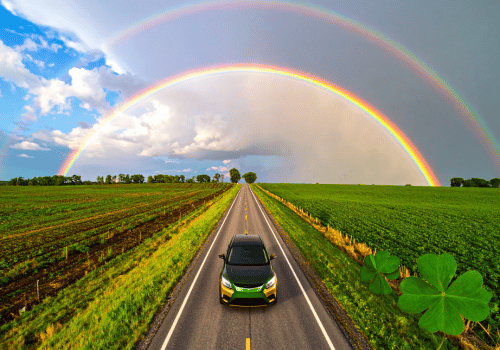 Car driving down a rural road under a double rainbow with shamrock graphics in the corner.