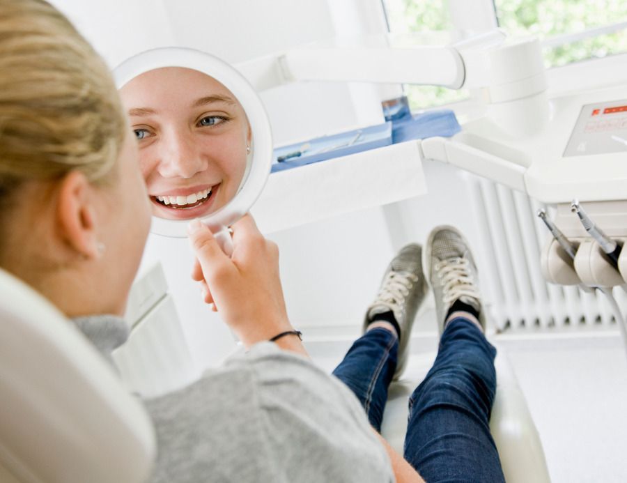 A woman is sitting in a dental chair looking at her smile in a mirror.