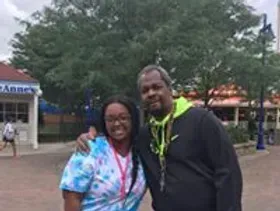 Two people smiling in front of a building and trees. Woman in blue, man in black jacket. Outdoor setting.