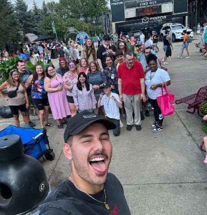 Man takes selfie with group in front of shops. Crowd of people outside; man sticking out tongue.