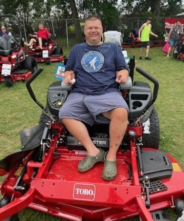 Man seated on a red Toro riding lawn mower. He's wearing a blue shirt and khaki shorts. Green grass and other mowers visible.