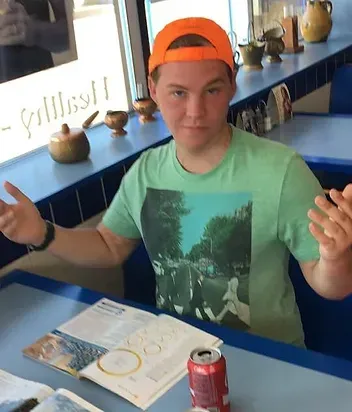 Person in green shirt and orange cap shrugs, seated at a blue table in a diner.