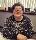 Woman in glasses, patterned sweater, smiling, seated behind a desk, with photos and a small sign visible in the background.