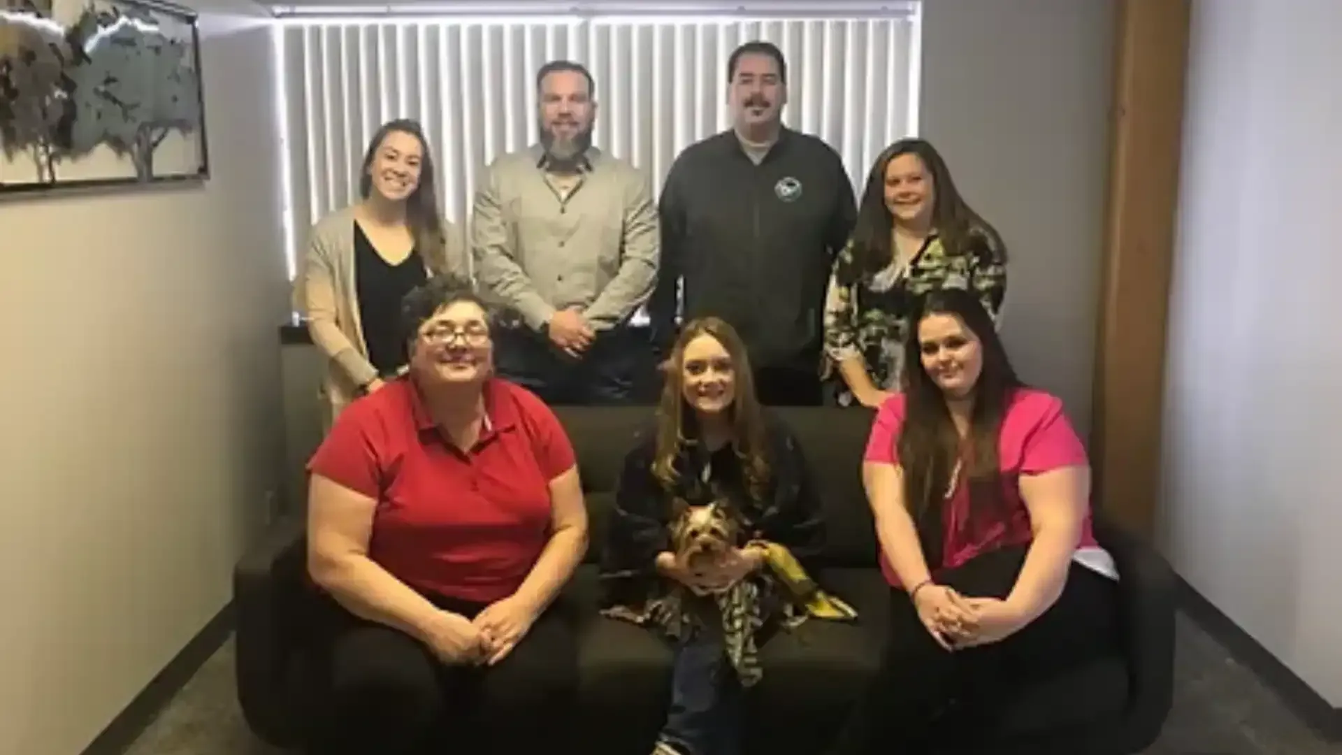 Group of seven people posing for a photo in an office setting, with a dog seated on a couch.