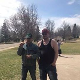 Two men standing outside near a road. One is smoking, the other wearing a tank top. Sunny day.