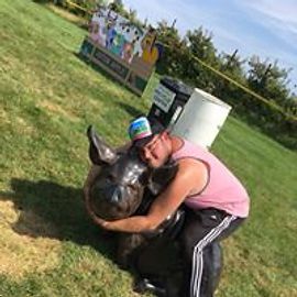 Person hugging a bronze pig statue outdoors, wearing a pink shirt and black pants.