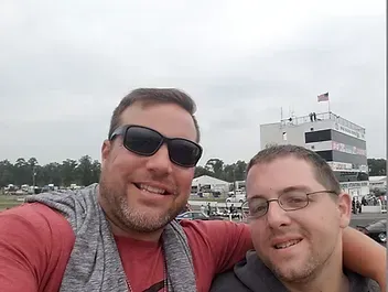 Two men smiling at a racetrack, one with sunglasses and a gray scarf. A tall control tower in the background.