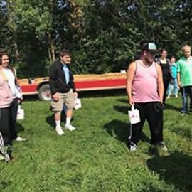 People standing in a grassy area near a trailer. One man wears a pink tank top and black pants. Others hold white bags.