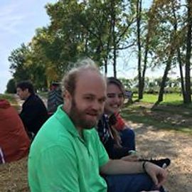 Man in green shirt smiles, sitting next to a woman; trees in background.