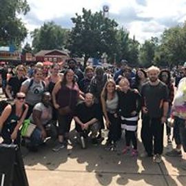 Large group of people outdoors, fair setting, some smiling, trees in background.