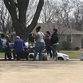 People gathered around picnic table, car parked nearby, house in background.