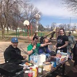 Group at picnic table in park, grilling food. People are seated and standing nearby.