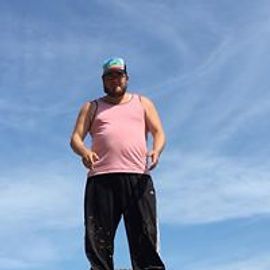 Man in pink tank top and black pants stands against a blue sky, wearing a hat.