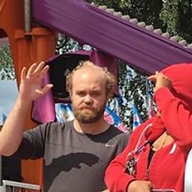 Man in gray shirt waves in front of purple ride structure.