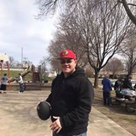 Man in red cap and black hoodie holding a ball outdoors at a park.
