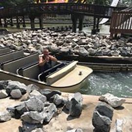 Man in a log flume boat on a water ride, surrounded by rocks and a bridge.