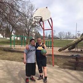 Two people stand near a basketball hoop. One holds a ball. The setting is a park.