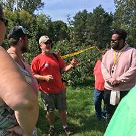Group of people outdoors in a grassy area, some talking near trees and a yellow object.