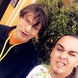 Two people smiling outdoors, one wearing glasses and a black jacket, the other with tattoos.