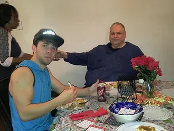 A man gestures toward another man at a dining table. A woman stands in the background. Flowers and food visible.
