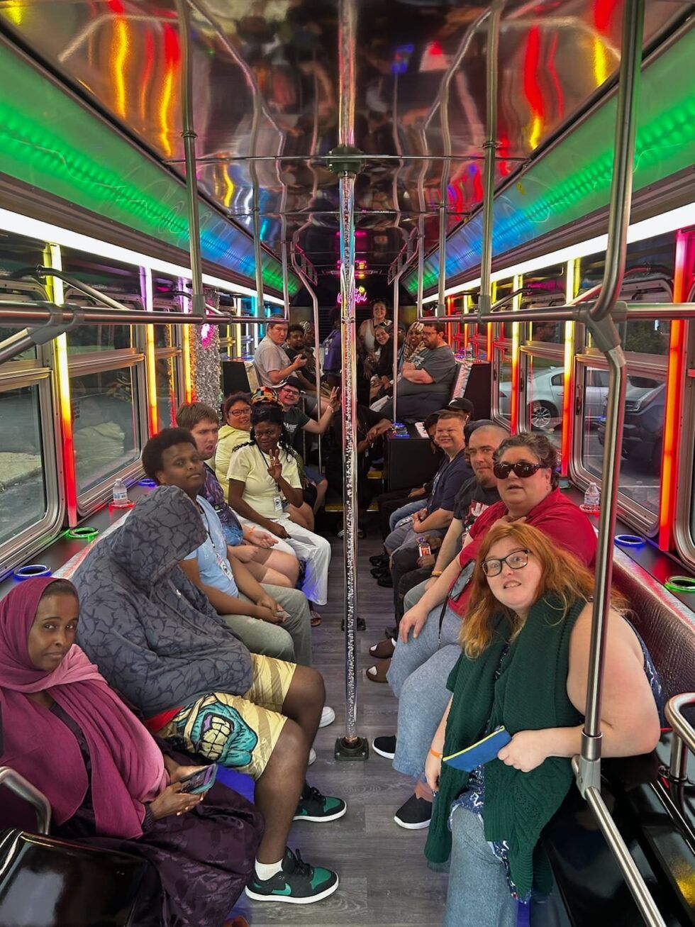 People seated inside a brightly lit vehicle, possibly a bus or trolley, with silver railings and colorful lights overhead.