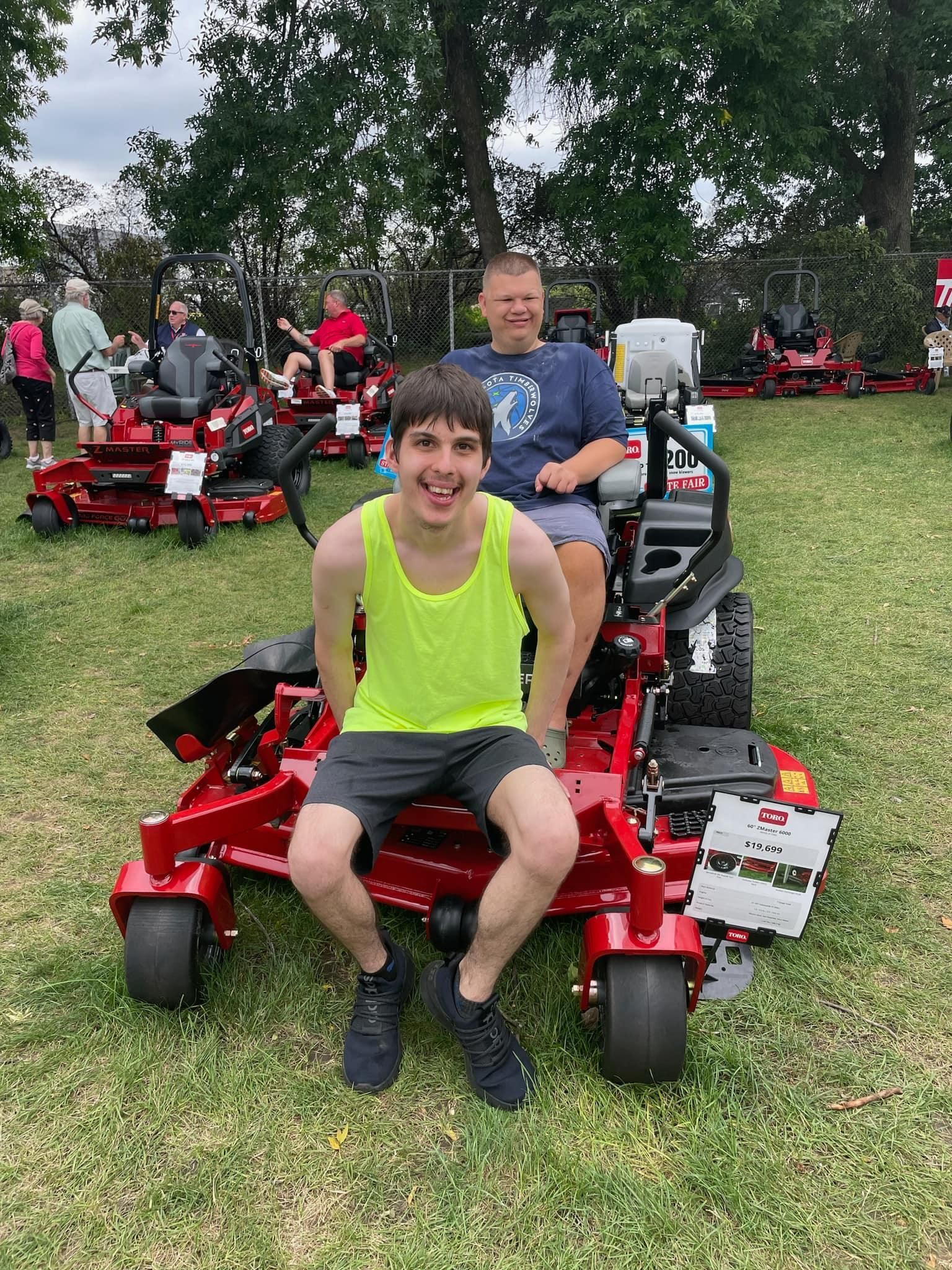 Two men smiling on a red lawnmower on a lawn. Other people and mowers in the background.