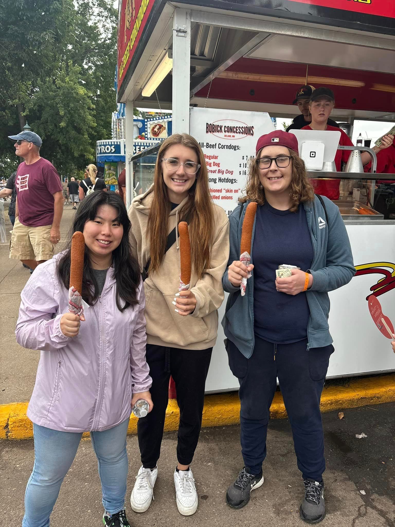 Three people hold corn dogs in front of a food stand.
