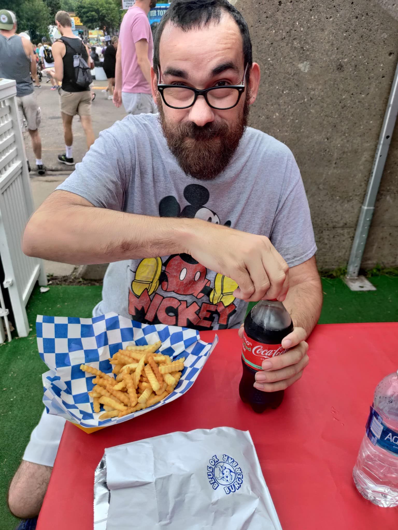 Man with glasses and beard opening a Coke bottle at an outdoor table with fries.